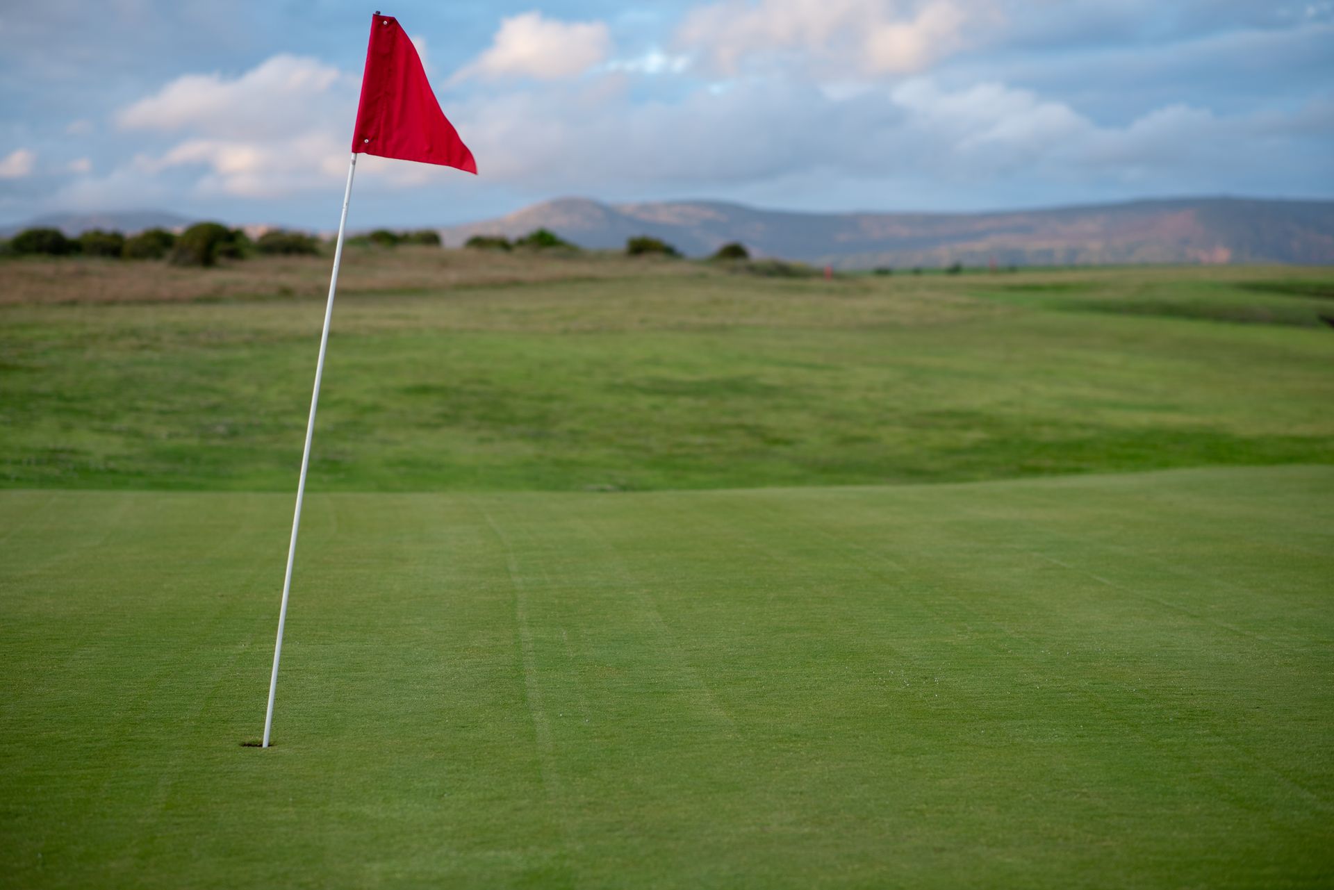 A golf green with a red flag on a sunny day, freshly mowed lawn with rolling hills in the background